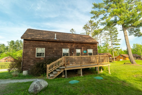 rustic cabin with a wooden deck surrounded by green trees and lawn