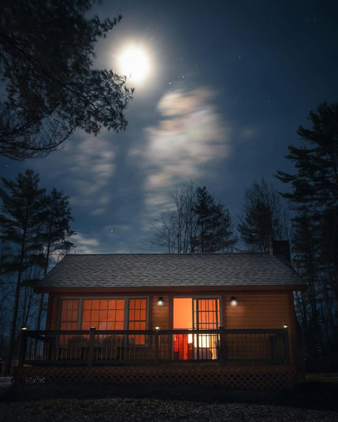 Cozy cabin with warm interior lighting under a bright moonlit sky.