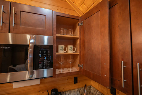 Open cabinet with glassware and mugs next to a stainless steel microwave in a rustic kitchen.
