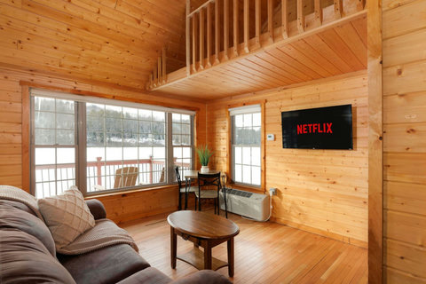 Cozy cabin living room with large windows overlooking a snowy landscape and a wall-mounted television.
