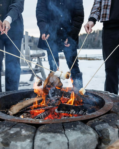 Guests roasting marshmallows around a stone fire pit in a snowy outdoor setting.