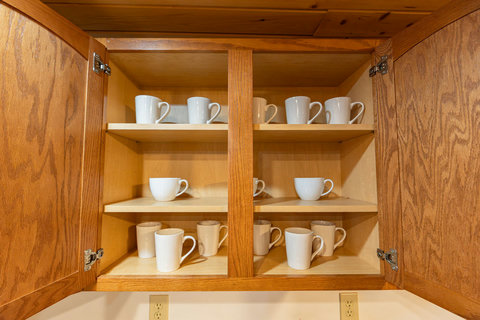 Wooden kitchen cabinet neatly stocked with white mugs for guest use.