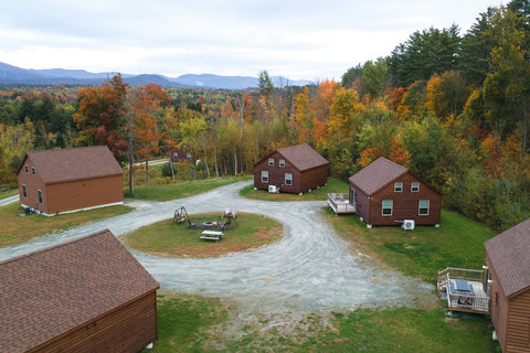Rustic cabins arranged around a central fire pit with fall foliage and mountain views in the background.