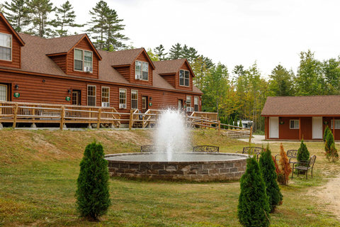 Rustic cabins surround a central stone fountain with seating in a wooded setting.