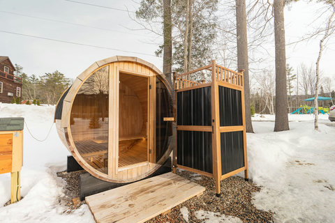 Outdoor barrel sauna with a private changing area surrounded by snow and trees.
