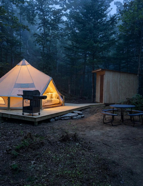Cozy glamping tent with outdoor grill and picnic table set in a forested area at dusk