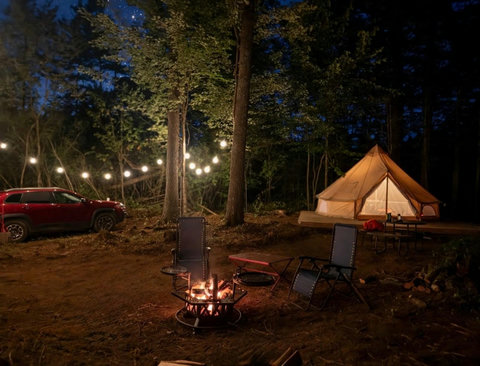 Cozy nighttime campsite with a lit fire pit, string lights, and a canvas tent in the woods.