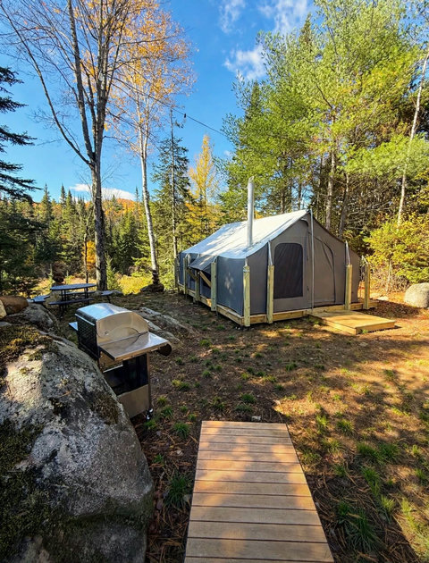 Rustic tent cabin nestled in a forest setting with a wooden deck and nearby picnic area.