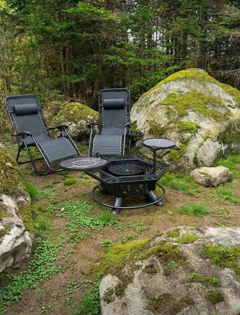 Outdoor seating area with lounge chairs, fire pit, and grill surrounded by forest and large rocks.