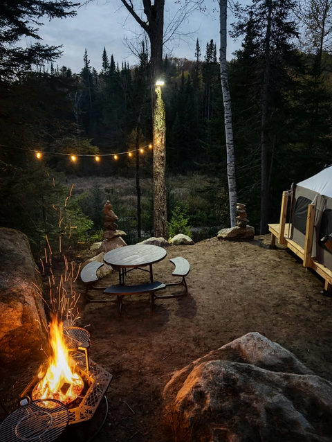 Outdoor picnic area surrounded by forest with a stone cairn and string lights for a peaceful retreat.